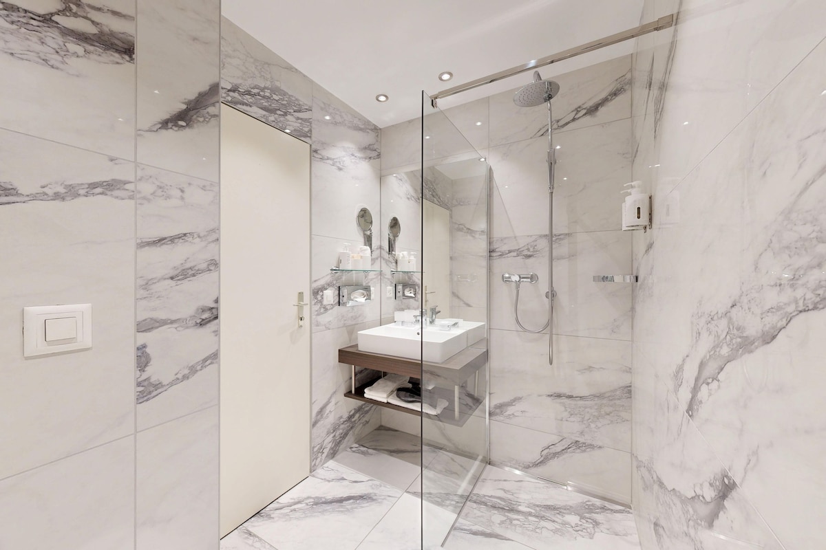 Un baño moderno con ducha envuelta en vidrio, paredes de mármol y lavamanos integrado.