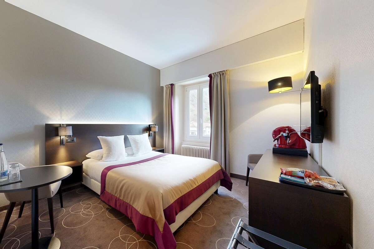 Une chambre d’hôtel comprenant un lit, une table de chevet, une lampe et un petit bureau avec une chaise.