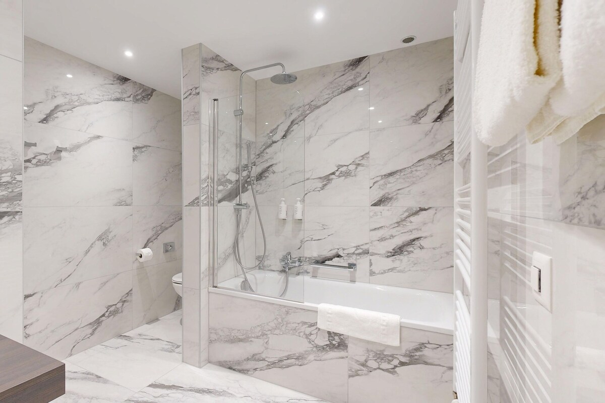 Une salle de bain moderne dotée d’une douche en verre, de murs en marbre et d’un meuble-lavabo blanc avec un miroir.