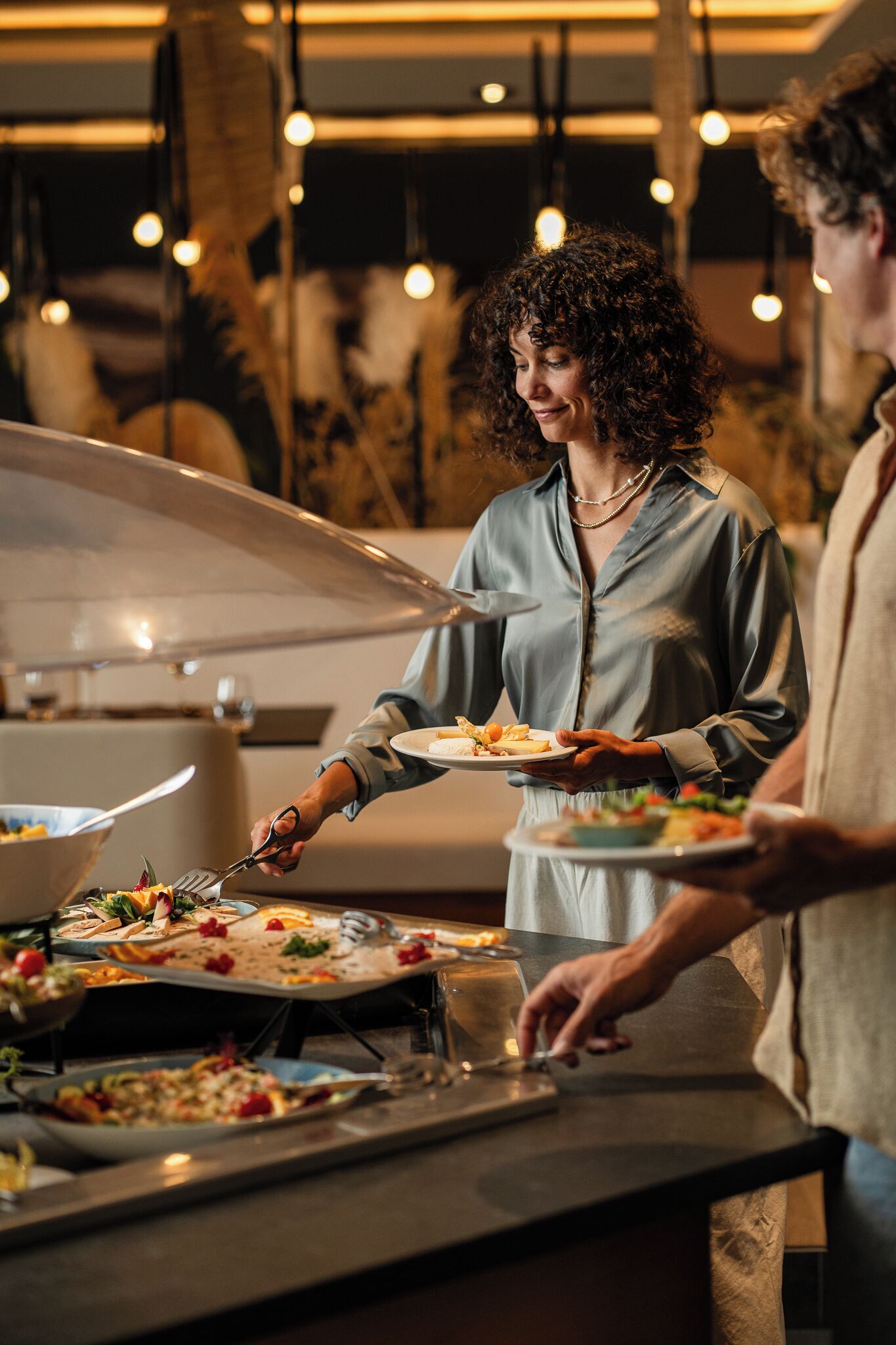 Een buffetopstelling met diverse gerechten, een persoon die eten opschept en een modern interieur met hangende lampen.