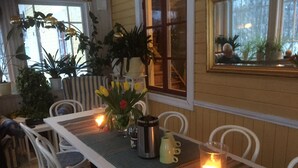 Un comedor con una mesa dispuesta para dos personas, rodeada de sillas blancas. Hay plantas en macetas, una vela y un termo sobre la mesa.