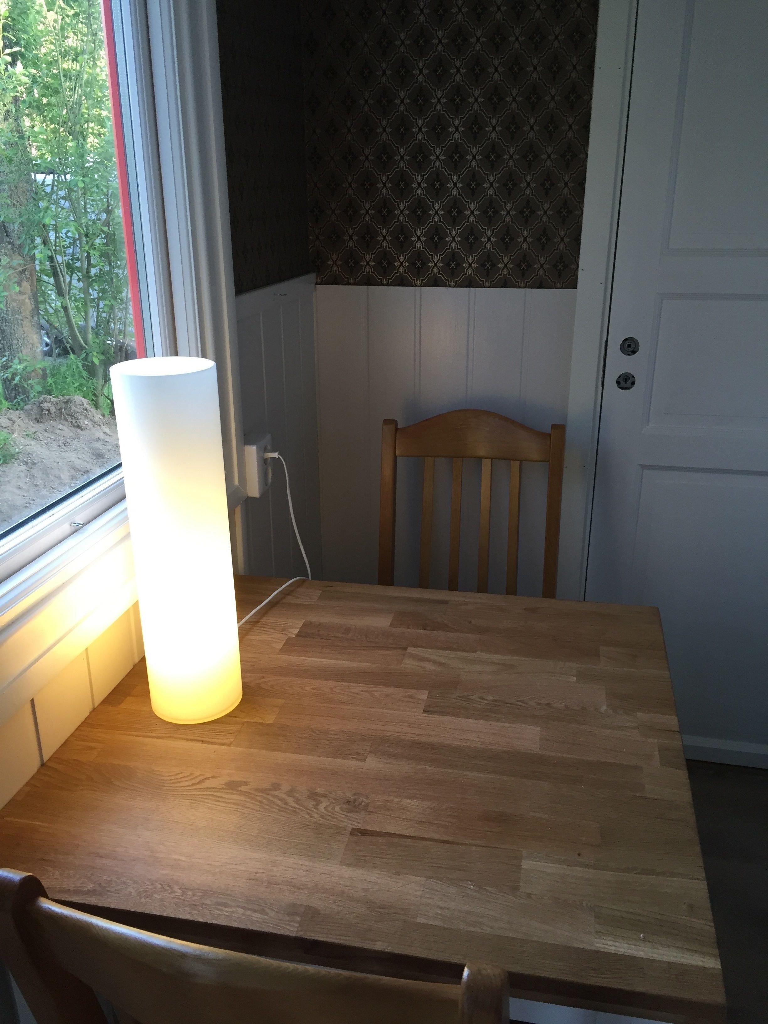 Una mesa de comedor de madera con una lámpara, una silla, una ventana con cortinas y un papel tapiz estampado.
