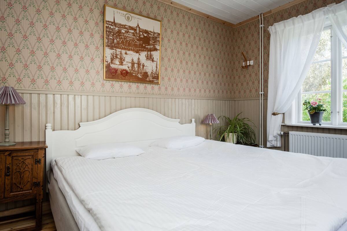 Un dormitorio con una cama grande, una mesita de noche, una lámpara de pared, una ventana con cortinas y una planta en maceta.