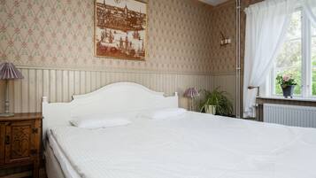 A bedroom with a large bed, a nightstand, a wall-mounted lamp, a window with curtains, and a potted plant.