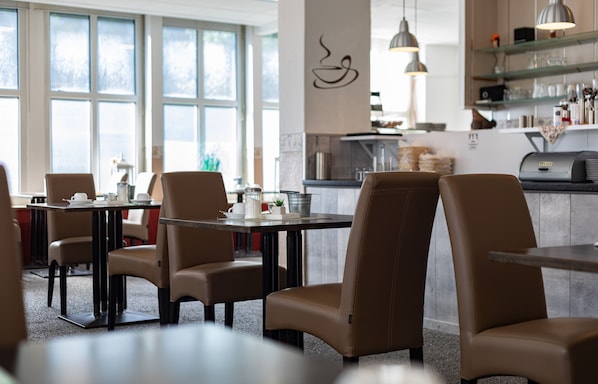 A modern restaurant interior with high tables and chairs, large windows, and a visible kitchen area. - Stadt-gut-Hotel Großer Kurfürst (Emden)