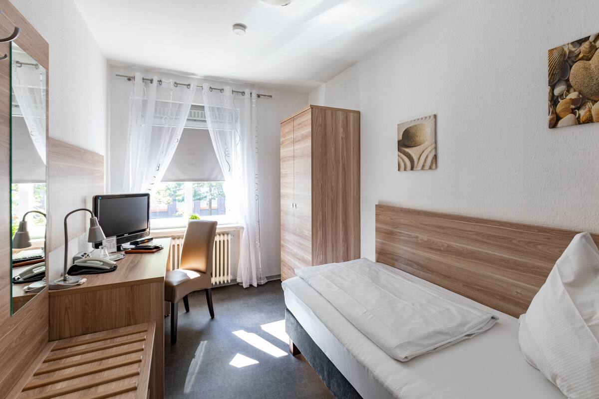 A hotel room with a bed, a desk with a computer, a chair, a radiator, and a window with sheer curtains.