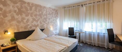 Una habitación de hotel moderna con una cama grande, una mesita de noche con lámpara, una pequeña mesa con silla y una ventana con cortinas translúcidas.