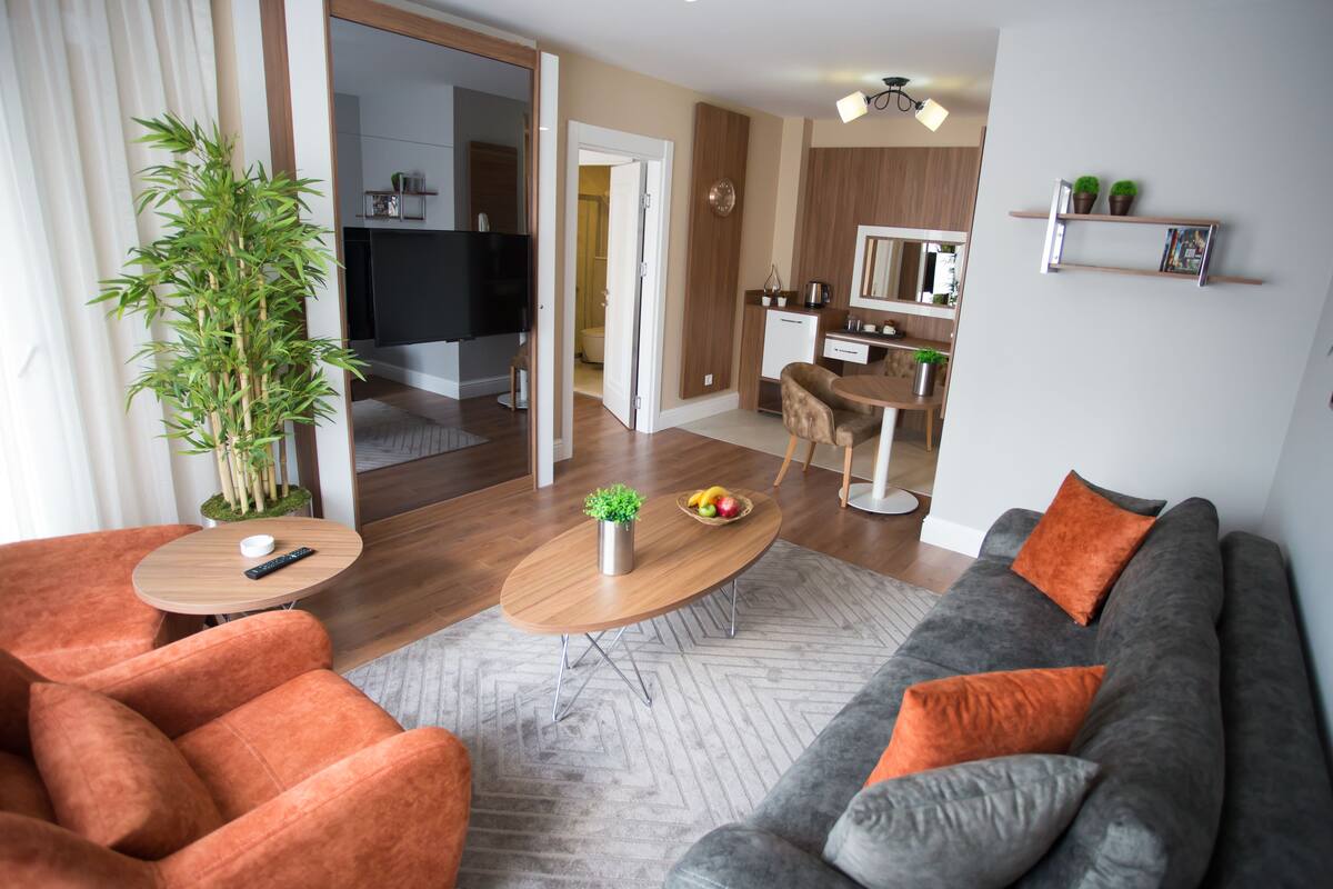 A modern living room with a sofa, armchair, coffee table, and a television. There is a kitchen area visible through an open door.