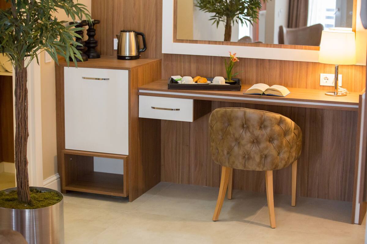 A modern hotel room with a wooden desk, a plush chair, a small plant, a tray with fruits and a book, and a coffee maker.