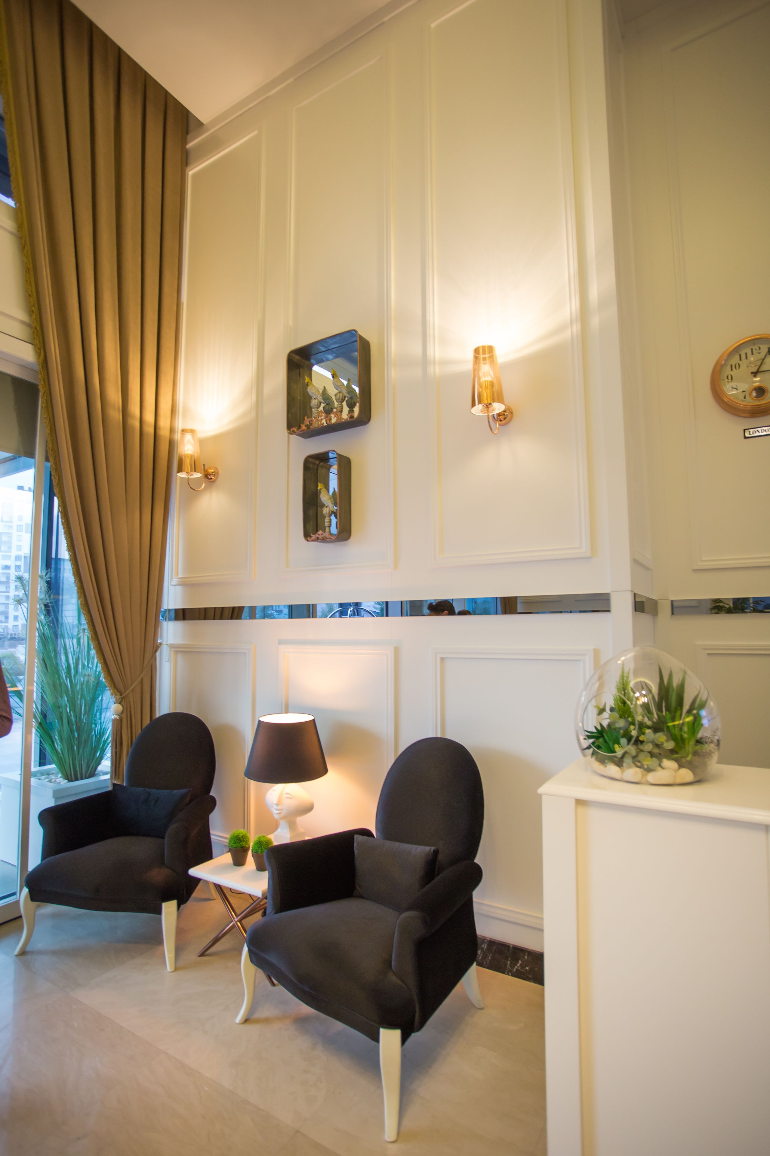 Eine Lobby mit zwei schwarzen Sesseln, einem kleinen Tisch mit Lampe, einer Topfpflanze und einem dekorativen Spiegel an der Wand.
