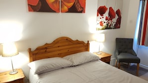A neatly made bed with a patterned bedspread, two bedside lamps, a wooden nightstand, a grey upholstered chair, and two wall-mounted paintings of red flowers. - Chelford Guesthouse (Knutsford)