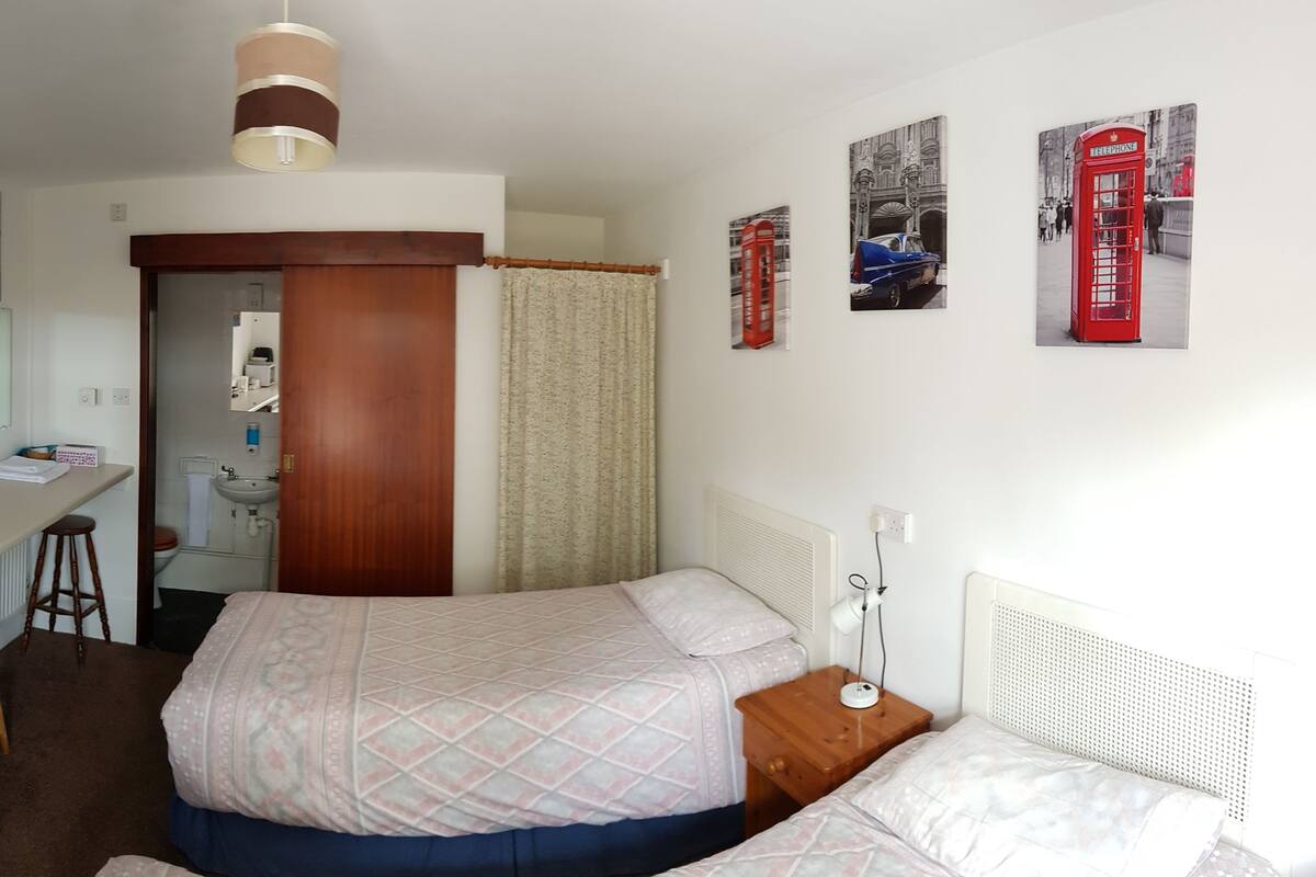 A hotel room with a kitchenette, two beds, a desk, and various wall decorations including a cityscape and a red telephone booth.