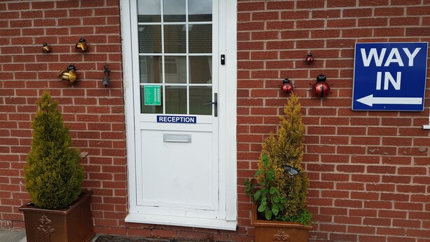 A reception area with a white door, a blue sign indicating 'WAY IN', and decorative elements like ladybug ornaments and a bell. - Chelford Guesthouse (Knutsford)