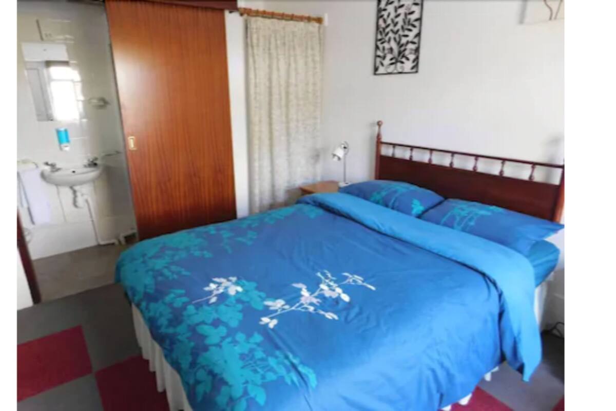 A bedroom with a bed, a nightstand, a lamp, a window with curtains, a wall-mounted artwork, and a bathroom with a sink and mirror.