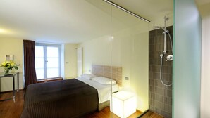 Habitación de hotel moderna con una cama grande, una mesita de noche con lámpara, zona de ducha y ventana con cortinas.