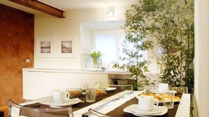 Un comedor con una mesa dispuesta para el desayuno, que incluye platos, tazas y un bol de cereales. Hay una planta grande al lado derecho de la mesa.