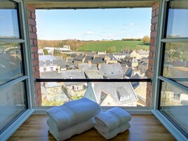 Quarto com vista para uma cidade através de uma janela, com piso de madeira e duas toalhas brancas enroladas.