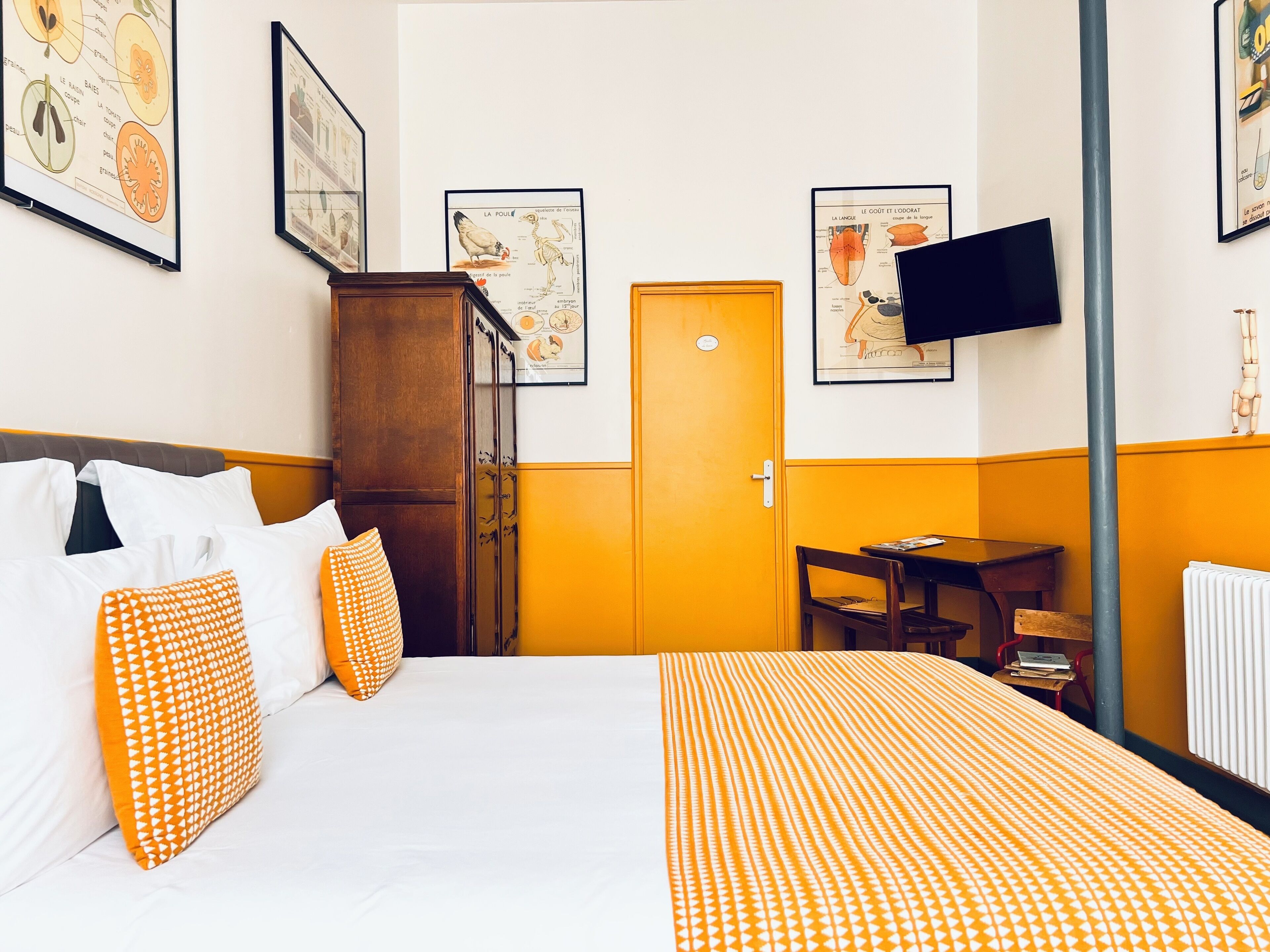 A hotel room with a bed, a wooden wardrobe, a desk, and a chair. There are framed educational posters on the walls.