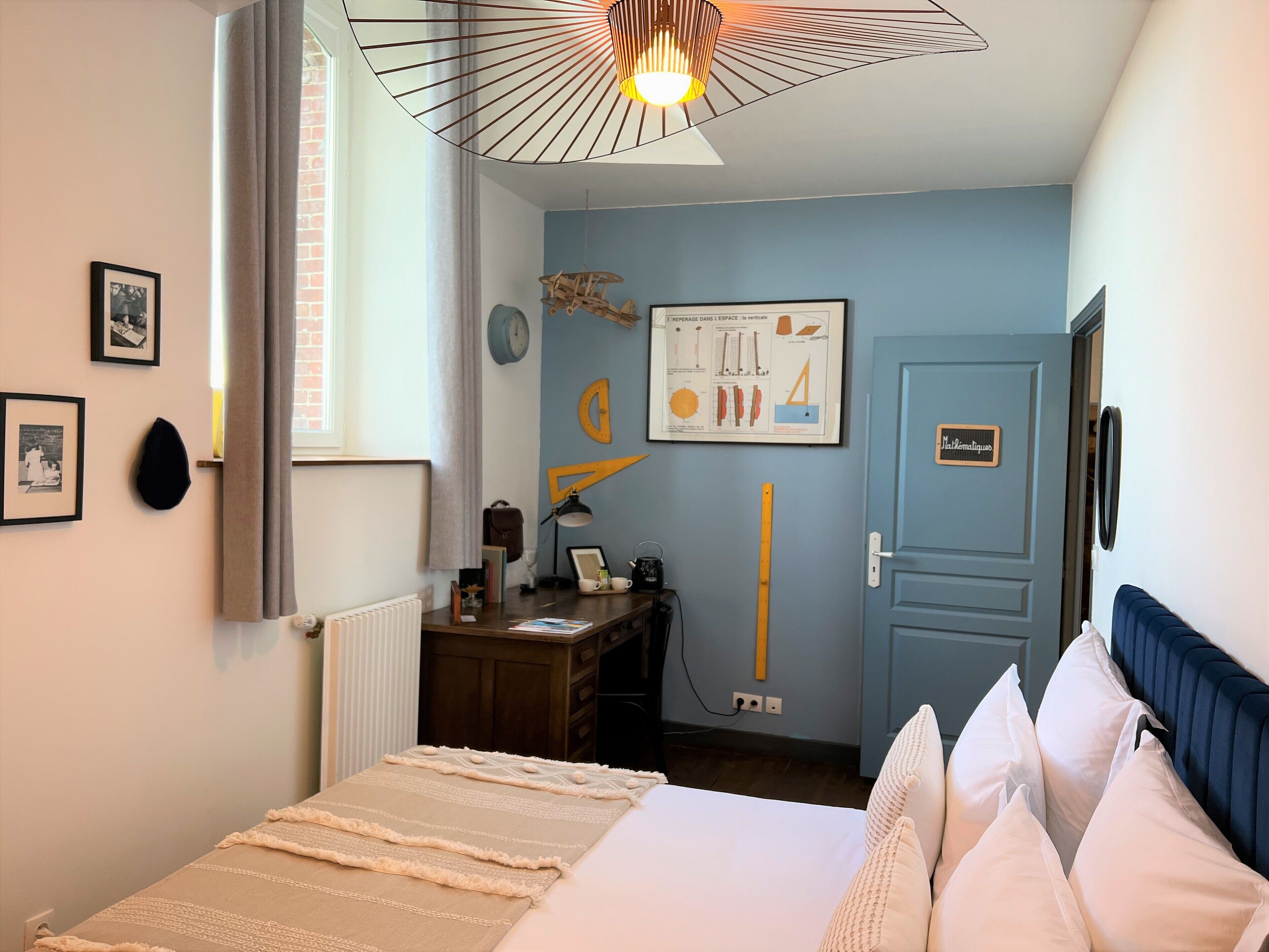 A hotel room with a bed, a desk, a chair, a lamp, a clock, and a door with a sign.