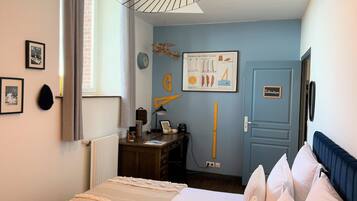 A hotel room with a bed, a desk, a chair, a lamp, a clock, and a door with a sign.