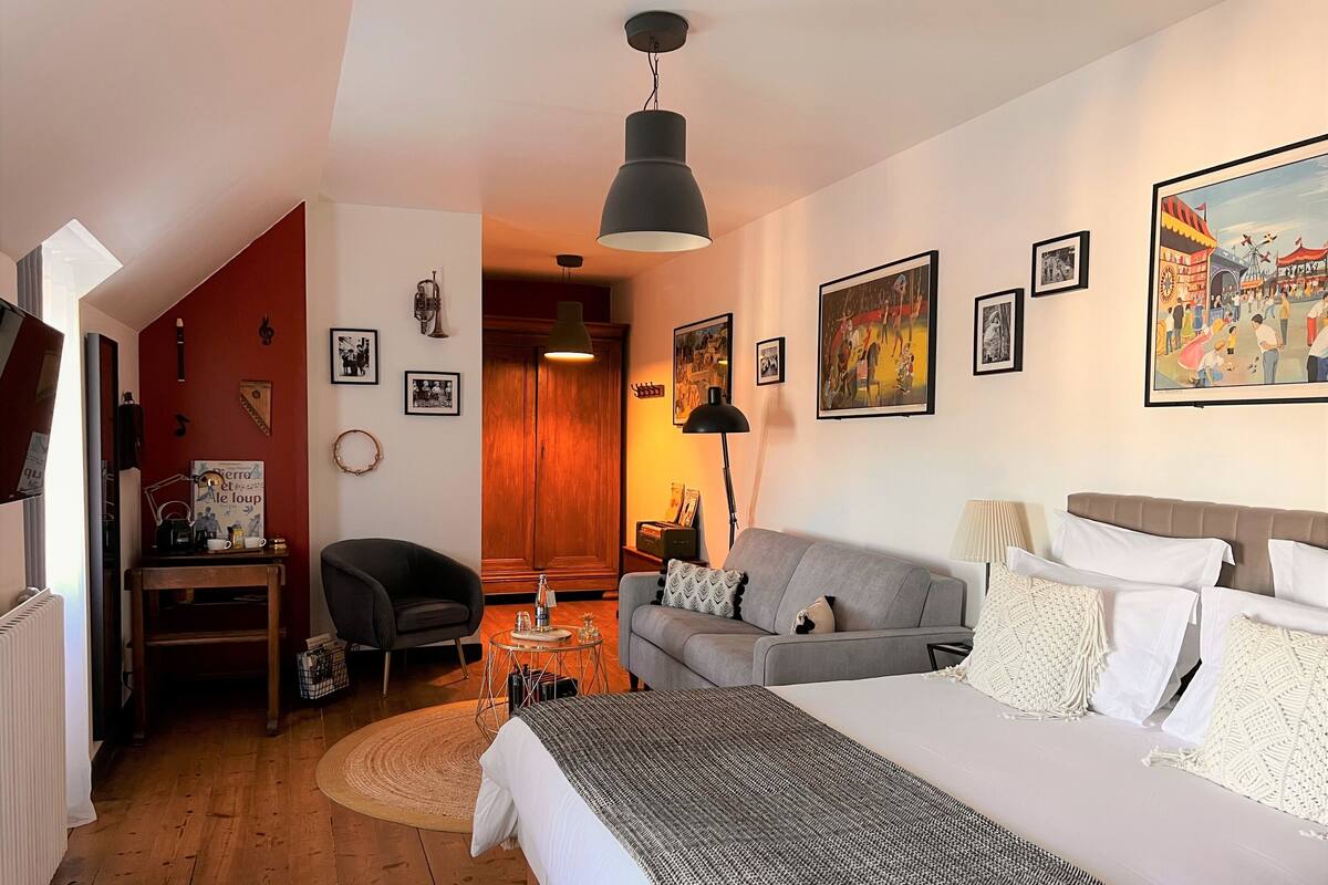 A hotel room with a bed, a sofa, a desk, and a chair. There are framed pictures on the walls and a lamp on the desk.