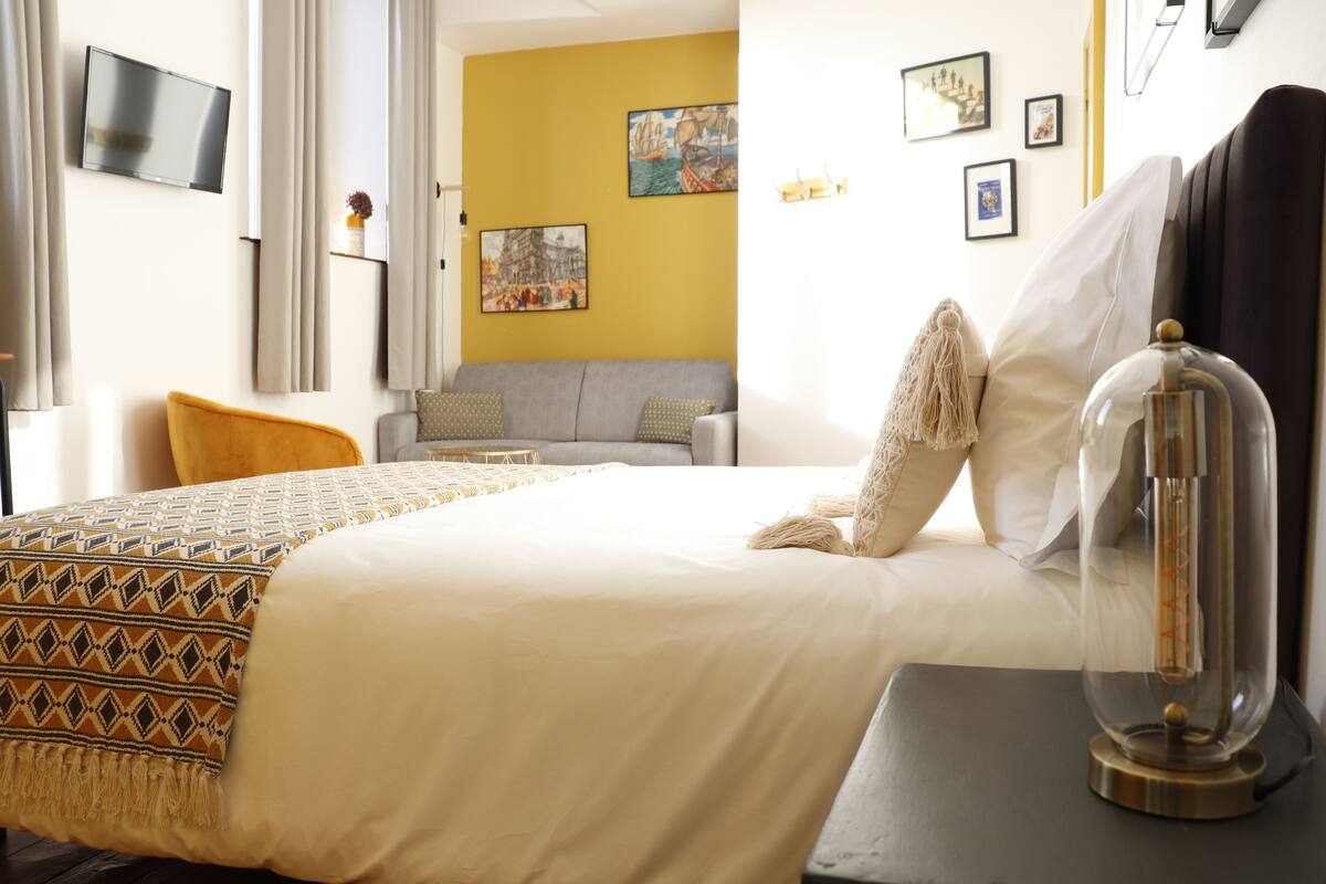A hotel room with a bed, a sofa, a TV, and artwork on the walls.