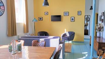 A modern living room with a sofa, chairs, a wooden dining table, and a TV mounted on the wall.