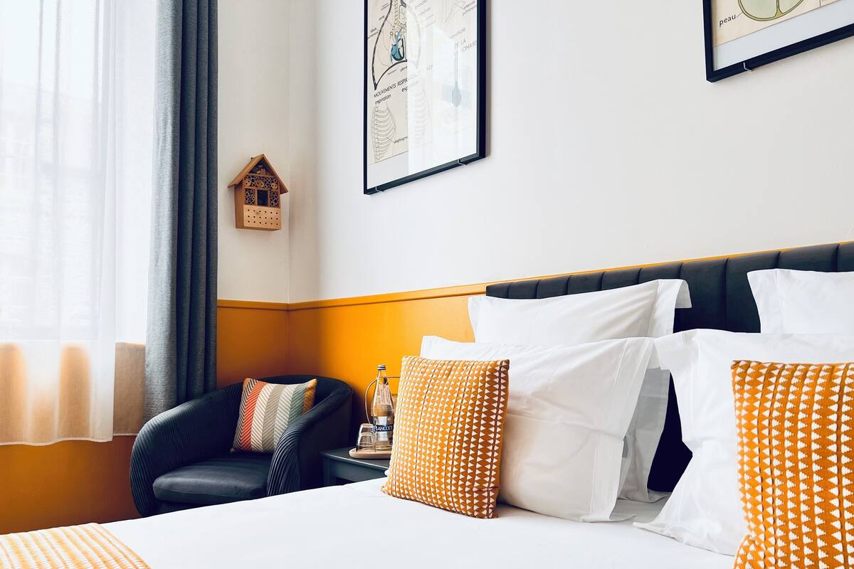 A hotel room with a bed, pillows, a chair, a nightstand, a bottle, and a framed map on the wall.