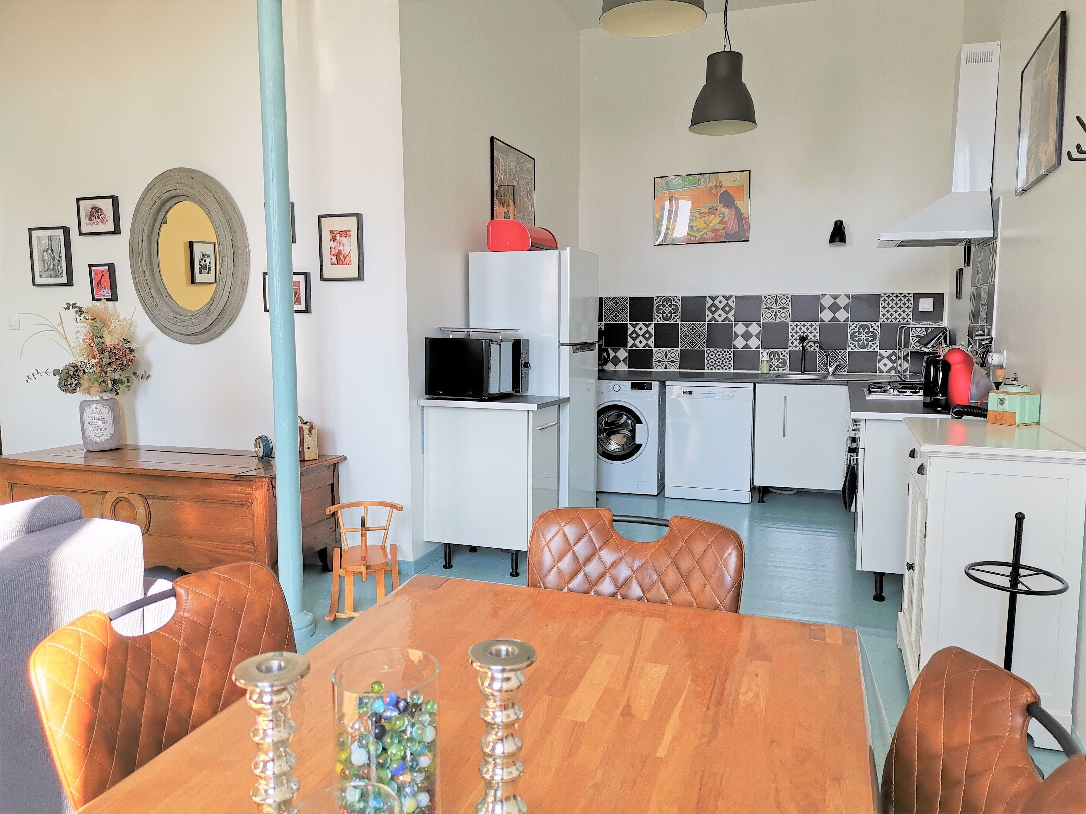 Cocina compacta con microondas, lavadora y un refrigerador pequeño. Hay una mesa de comedor con sillas y un florero con flores.
