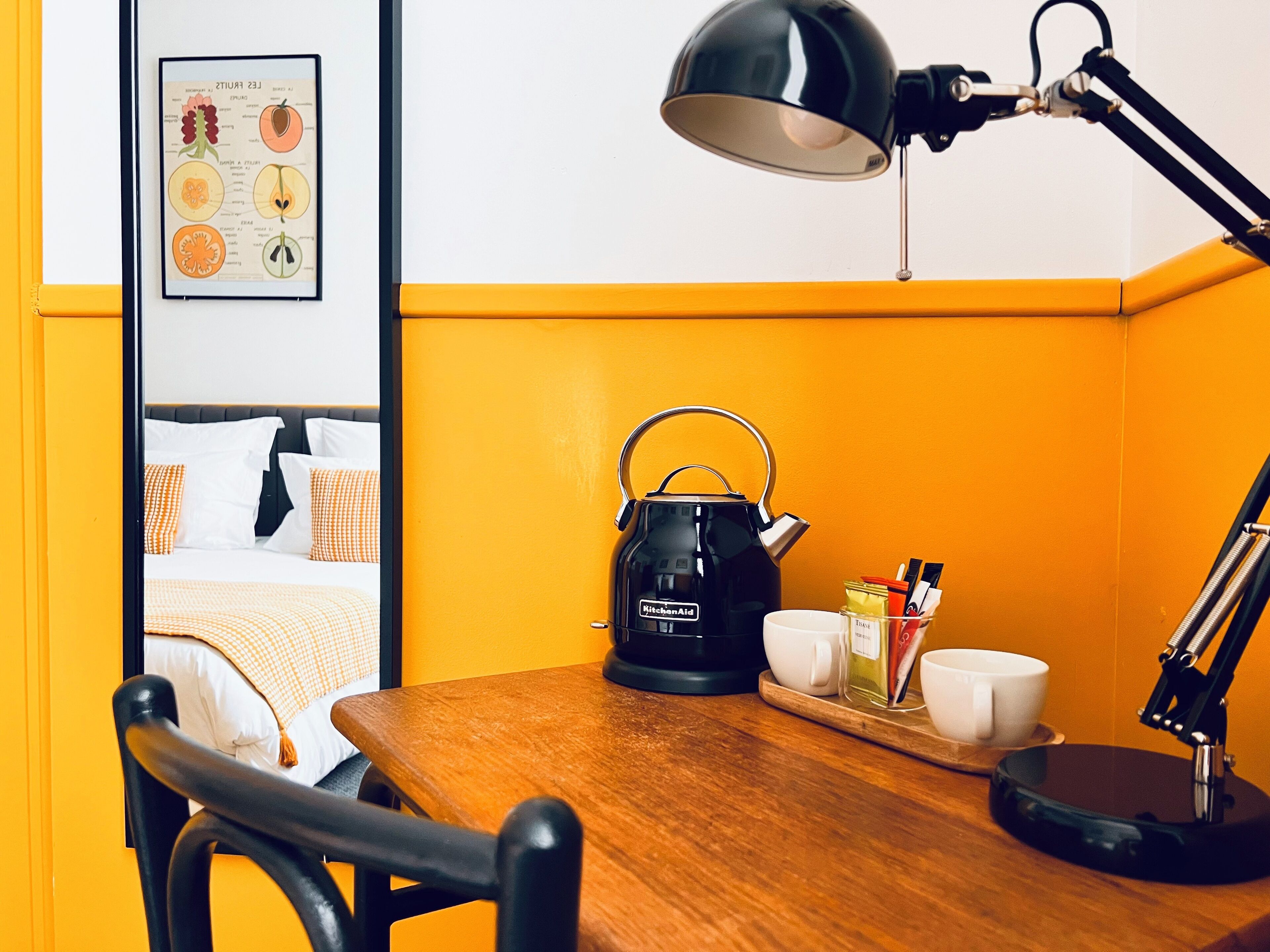 Una habitación de hotel con una mesa de madera, un hervidor eléctrico negro, una lámpara de escritorio e ilustración botánica enmarcada en la pared.