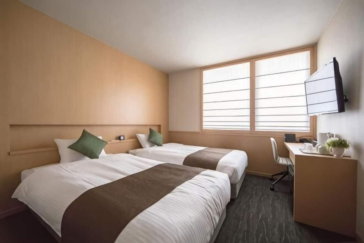 A hotel room with two beds, a desk, a chair, a TV, and a window.