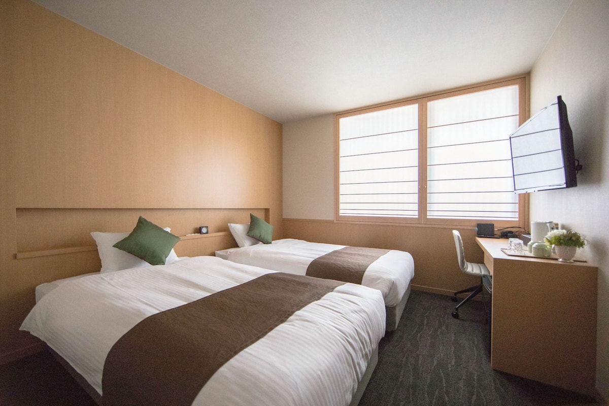A hotel room with two beds, a desk, a chair, a TV, and a window with blinds.