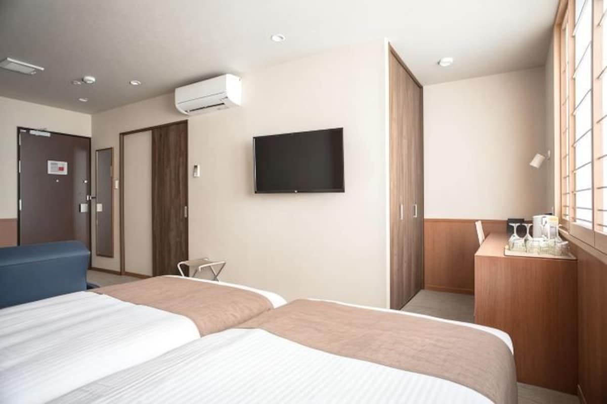 A hotel room with two beds, a TV, a desk with a chair, and a window with blinds.