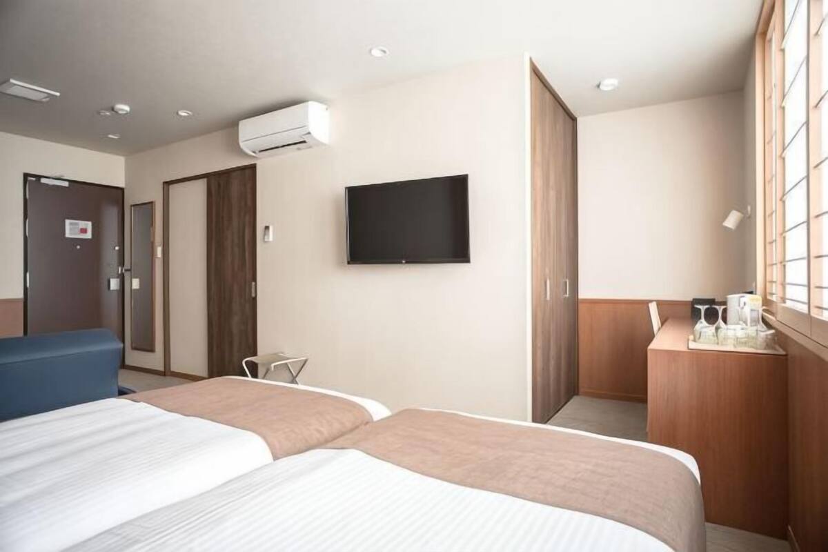 A hotel room with two beds, a TV, a desk with a chair, and a window with blinds.
