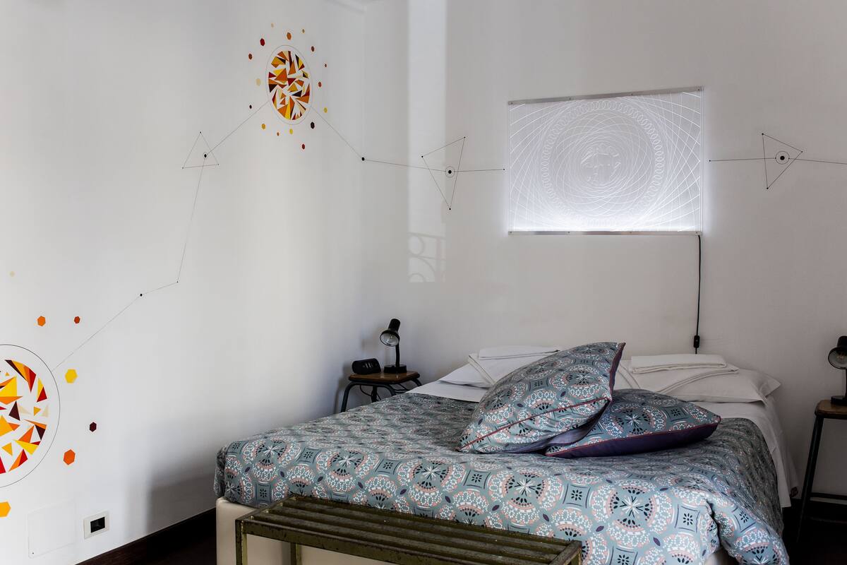 A bedroom with a patterned bedspread, a window with blinds, and abstract wall art.