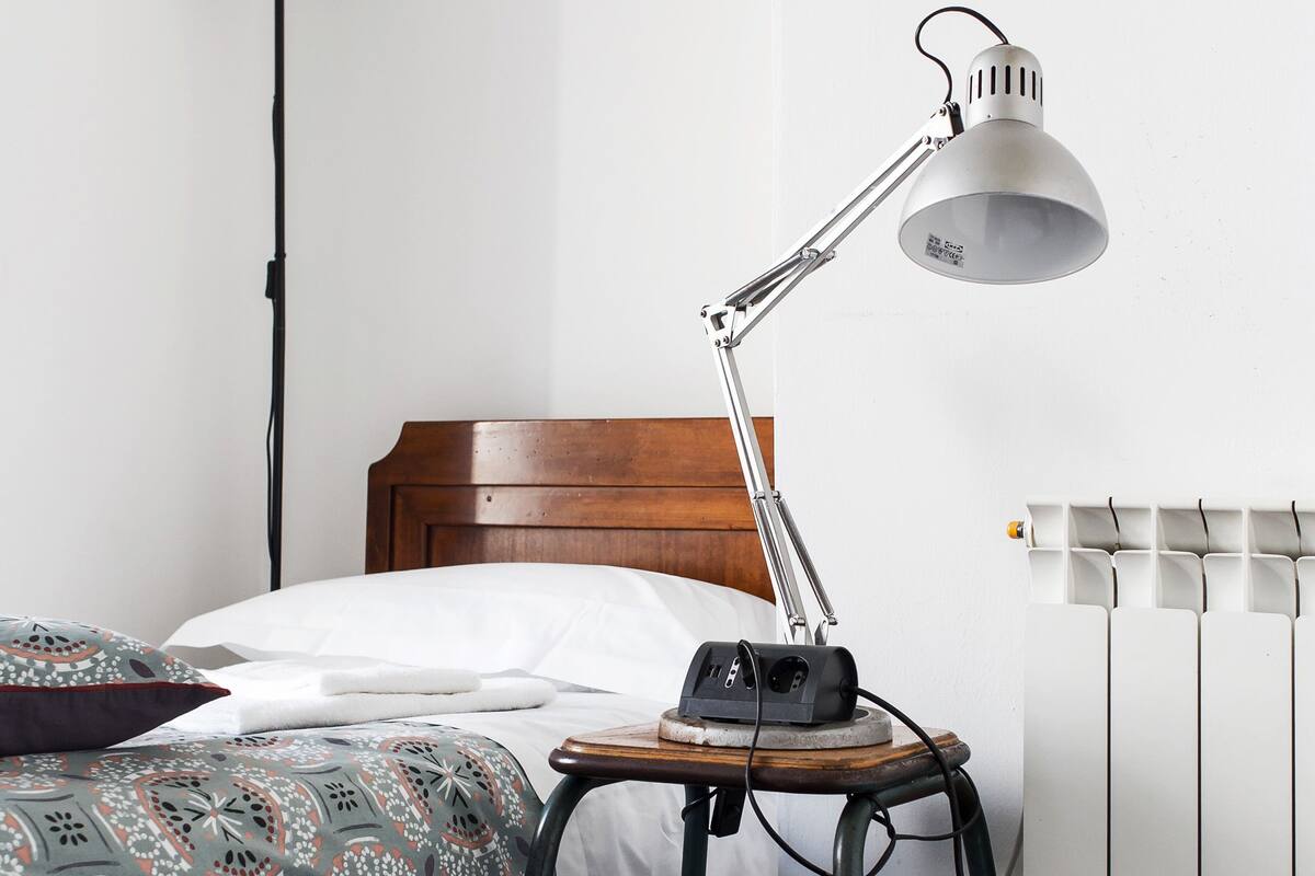 A neatly made bed with a patterned bedspread, a bedside lamp, a small table with a radio, and a radiator in a room with white walls and dark wooden flooring.