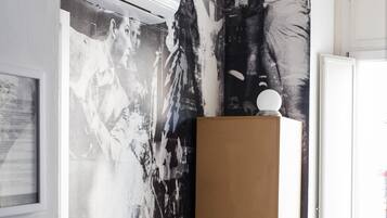A room with a large black and white mural on the wall, a wooden chair, a bed with patterned bedding, and an air conditioning unit mounted on the wall.