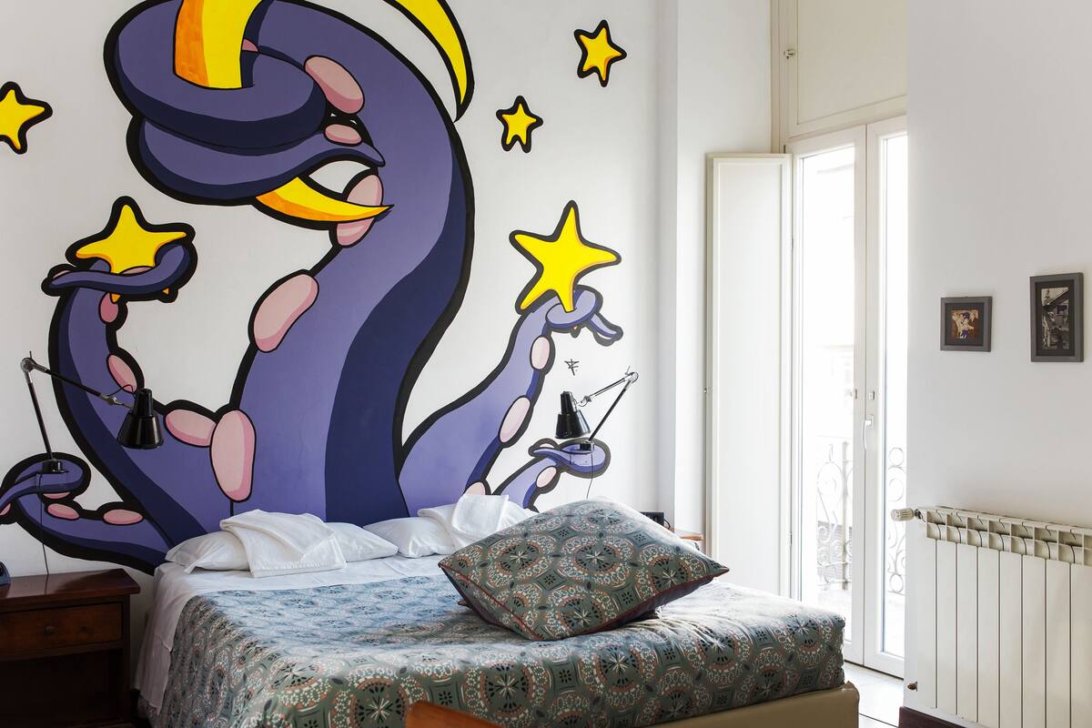 A bedroom with a bed, a nightstand, a wall mural, and a window.