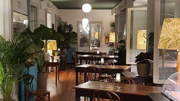 Restaurante com mesas e cadeiras de madeira, plantas em vasos e luminárias pendentes.