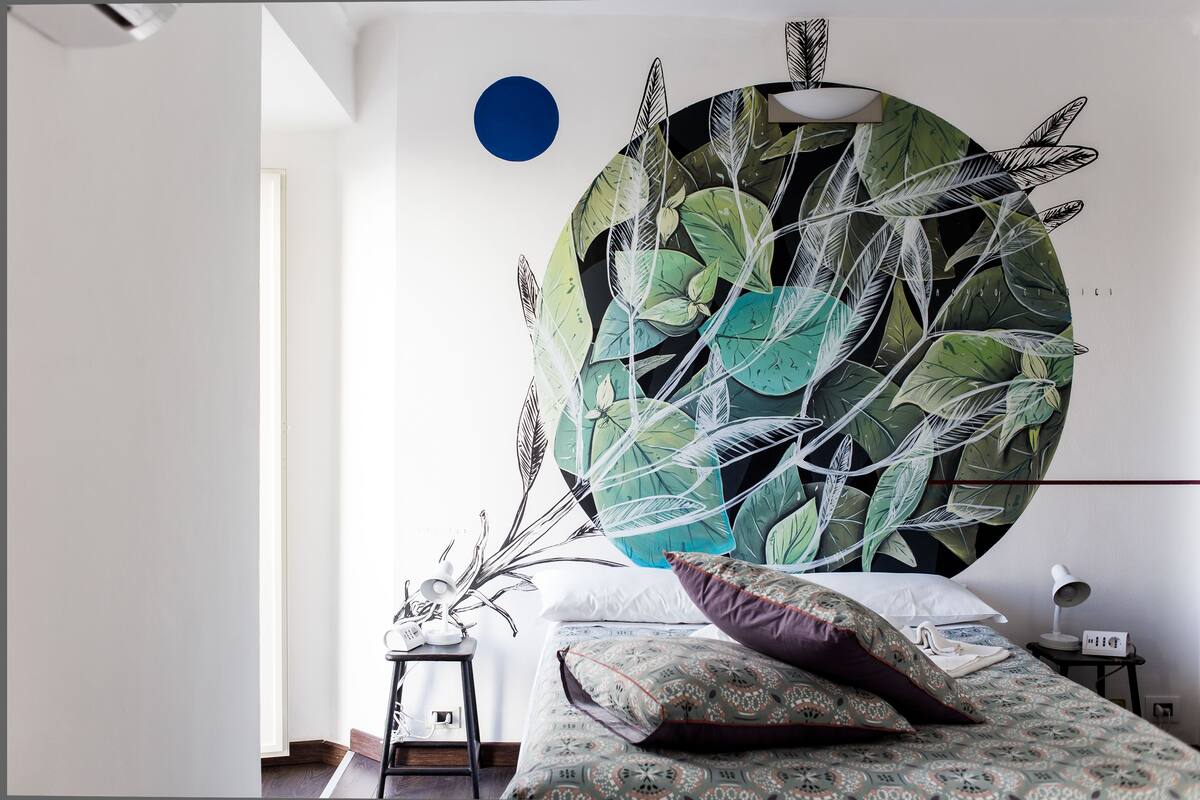 A bedroom with a large wall mural of tropical leaves, a bed with patterned bedding, a small side table with a lamp, and a wall-mounted light fixture.