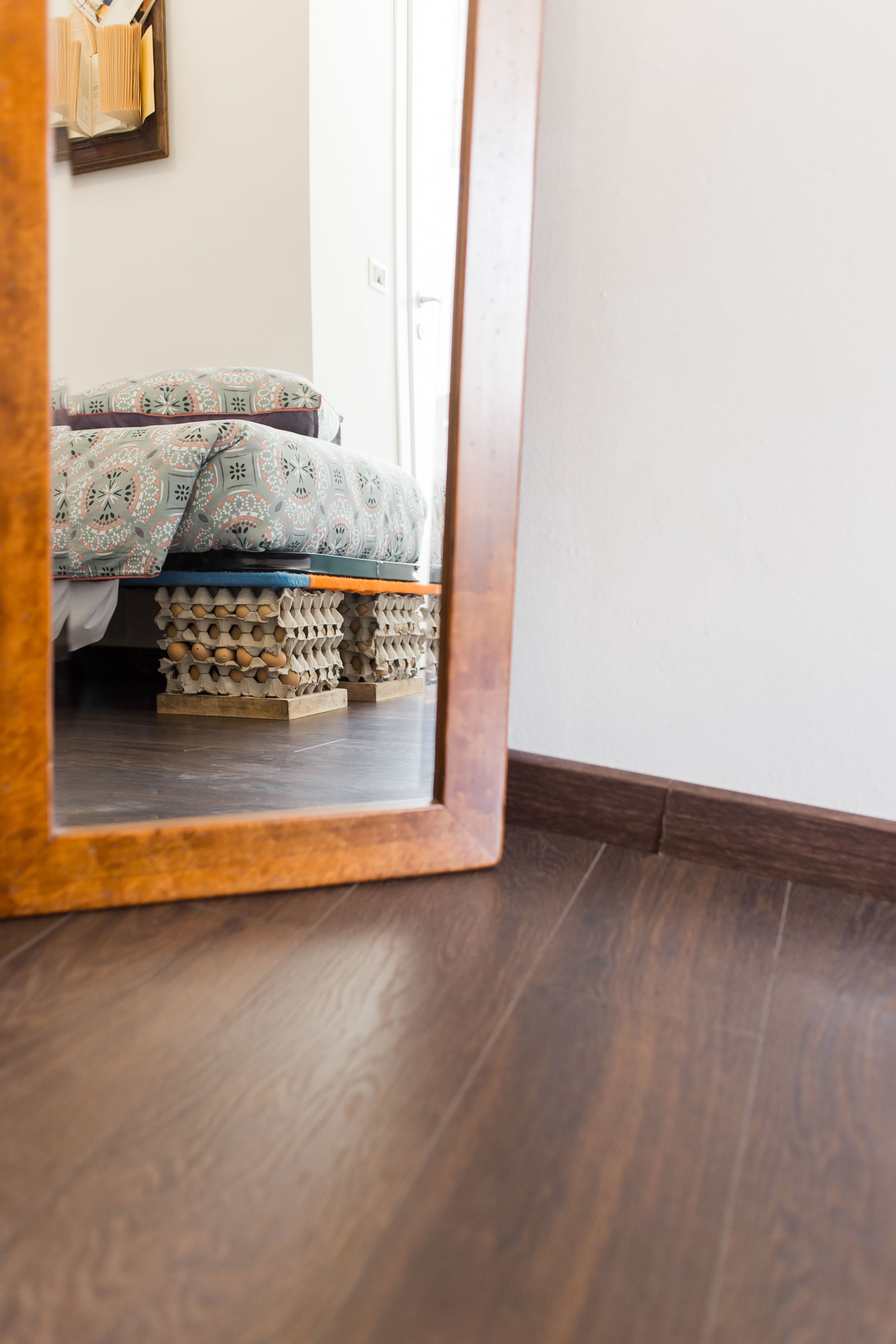 A wooden-framed mirror reflecting a bed with patterned bedding and a stack of egg cartons on the floor.