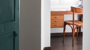 Quarto com escrivaninha de madeira, uma cadeira, janela com cortinas e porta azul-turquesa.