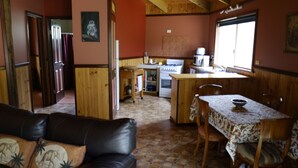 Uma sala de estar aconchegante com sofá de couro, mesa de jantar com cadeiras, área de cozinha com fogão e pia, e paredes e pisos de madeira.