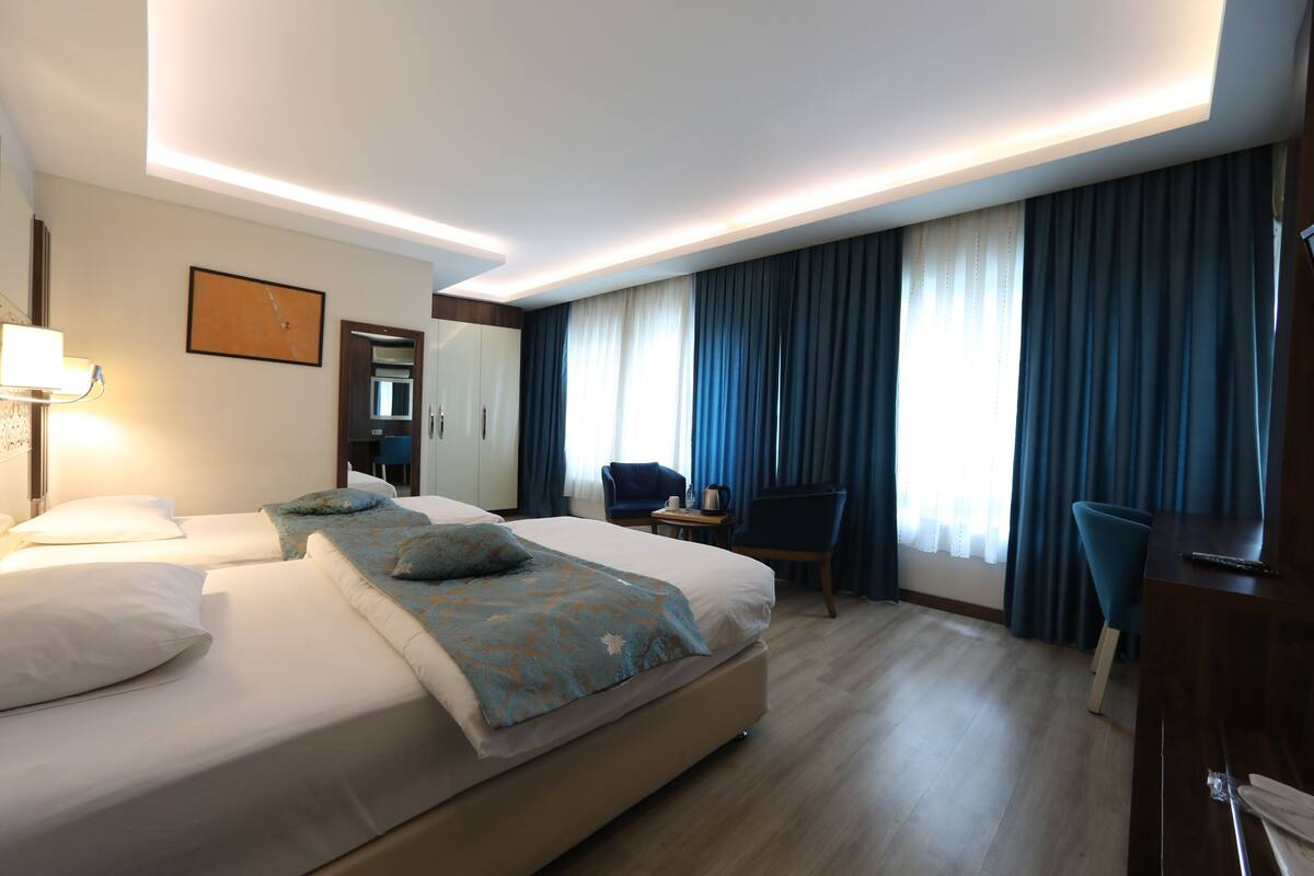 Quarto de hotel com duas camas, uma escrivaninha, uma cadeira e uma janela grande com cortinas.
