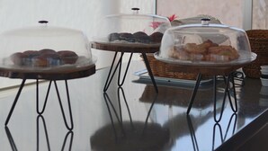 Una mesa de bufé con diferentes platos cubiertos, incluyendo pasteles y galletas, sobre una superficie reflectante, con una silla de mimbre al fondo.