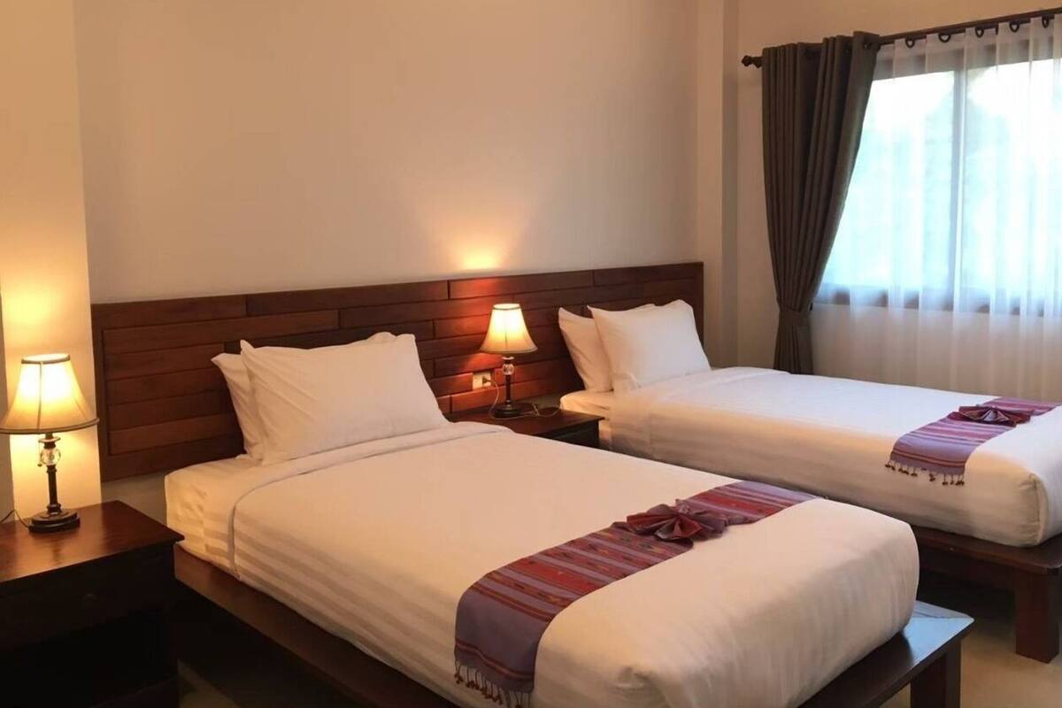A hotel room with two single beds, wooden headboards, bedside lamps, and a window with curtains.