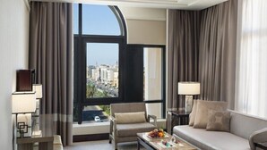 Una habitación de hotel moderna con un ventanal grande, un sofá, una silla, una mesita de centro con fruta y un vaso, y dos lámparas de noche.