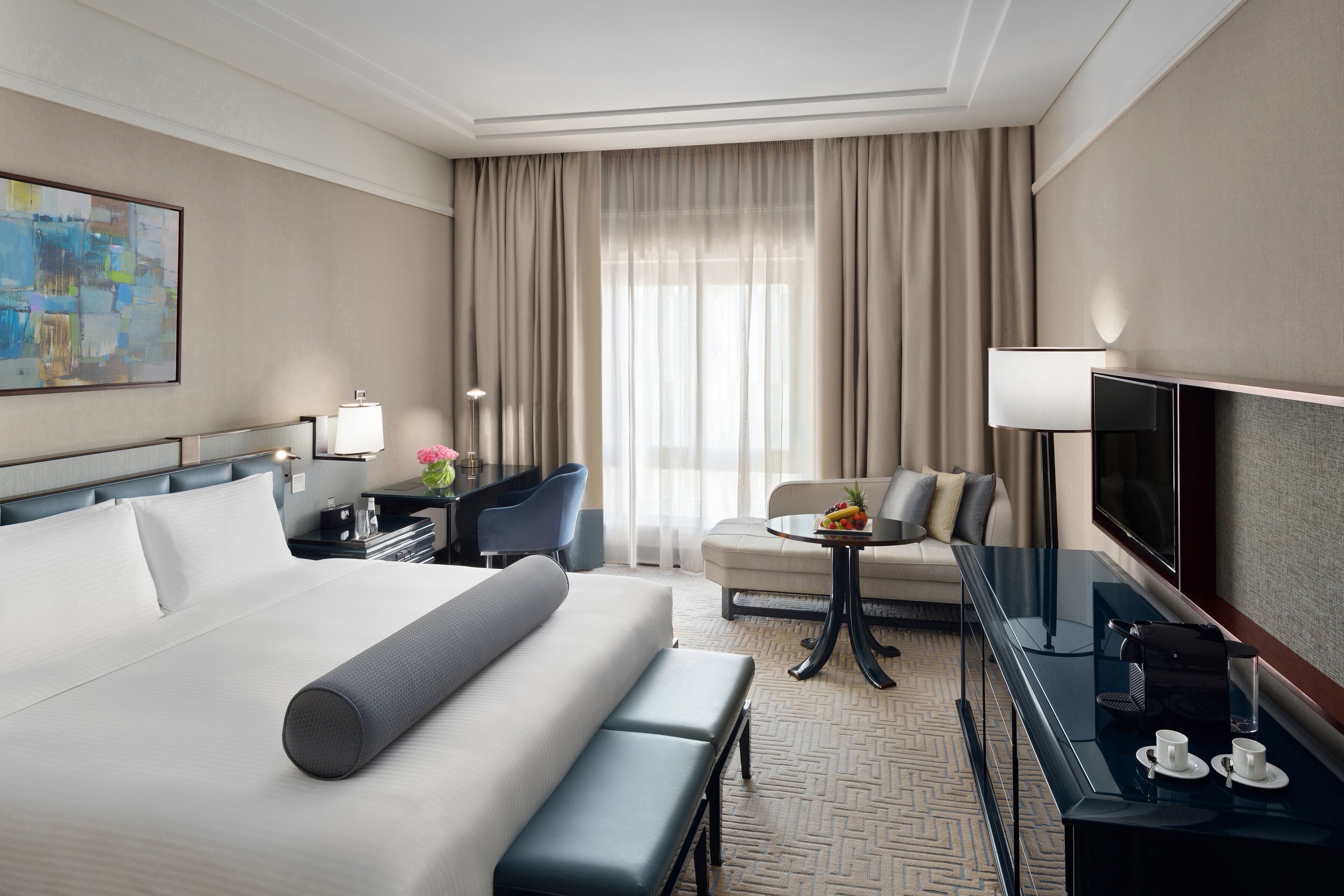 A hotel room with a large bed, a desk with a lamp, a chair, a sofa, a TV, and a coffee table with a fruit bowl.