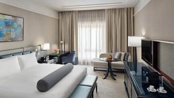 A hotel room with a large bed, a desk with a lamp, a chair, a sofa, a TV, and a coffee table with a fruit bowl.