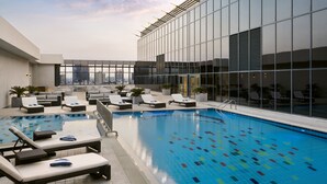 Una piscina en la azotea con sillones de descanso y un edificio de vidrio al fondo.